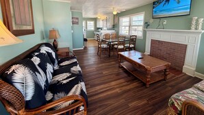 A living room with a sofa, dining table, chairs, a fireplace, and a kitchen in the background.