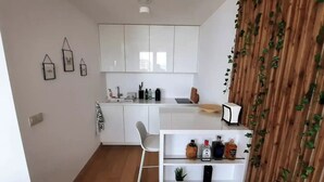 A compact kitchen with white cabinets, a sink, and a small dining area with a chair. There is a shelf with various bottles and a potted plant. - City view from 27th floor, free parking, keyless (Bratislavský kraj)