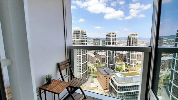 A balcony with a wooden chair and table, overlooking a cityscape with modern buildings. - City view from 27th floor, free parking, keyless (Bratislavský kraj)
