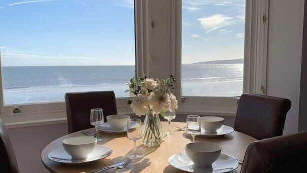 A dining table set for four with white bowls, plates, and glasses, and a vase of flowers. The table is positioned near large windows offering a view of the ocean. - Apartment 14, The Moorings  Beachfront & panoramic sea views (Filey)