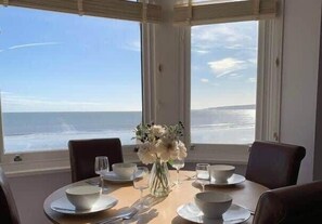 Una mesa de comedor lista para cuatro, con cuencos, platos y vasos blancos, y un jarrón con flores. La mesa está colocada cerca de grandes ventanales con vista al océano.