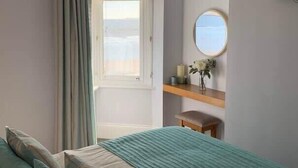 A bedroom with a bed, a window with curtains, a round mirror, a small table with a vase, and a view of the outside. - Apartment 14, The Moorings  Beachfront & panoramic sea views (Filey)