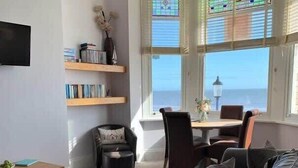 A living room with a view of the sea, a bookshelf, a round table, and a sofa. - Apartment 14, The Moorings  Beachfront & panoramic sea views (Filey)