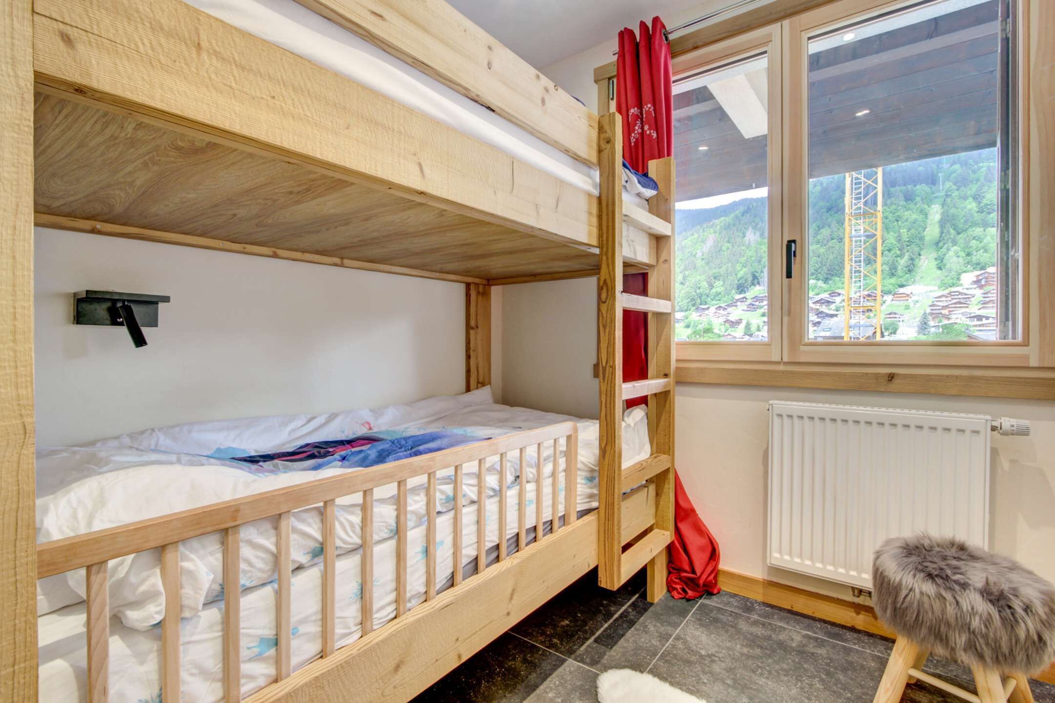 A bunk bed room with a window view of a forest and buildings, a radiator, and a small stool.
