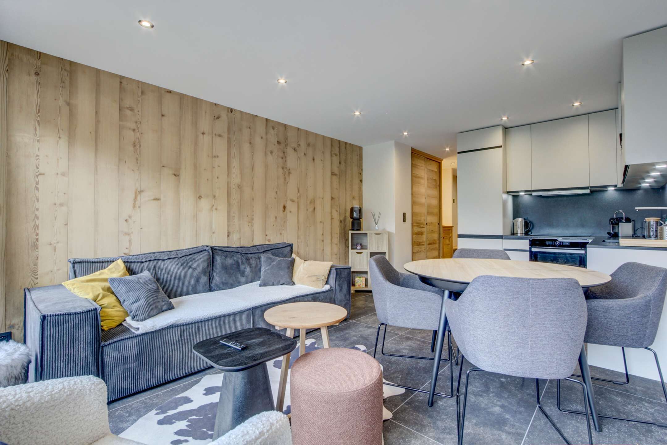 Una sala de estar moderna con sofá, mesa de centro y sillas, un área de cocina con electrodomésticos, y una pared de madera.
