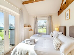 Uma cama de casal com roupa de cama branca e cabeceira, uma mesa de cabeceira com abajur, uma janela com cortinas e vista para uma paisagem verde.