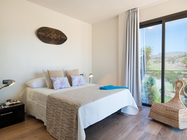 A bedroom with a bed, pillows, a bedside table with a lamp, a wall decoration, and a view of the outdoors through a large window.