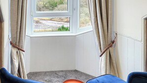A room with a bay window, beige curtains, a blue chair, and a view of a natural landscape. - 3 bedroom accommodation in Larkhall (Larkhall)