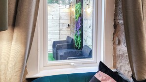 A room with a teal sofa, a window with curtains, and a view of an outdoor area with a wooden fence and greenery. - 3 bedroom accommodation in Larkhall (Larkhall)