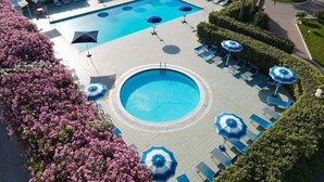 Una amplia piscina rectangular con una piscina circular más pequeña, rodeada de sillas de descanso y sombrillas, con jardines bien cuidados y un cielo despejado.