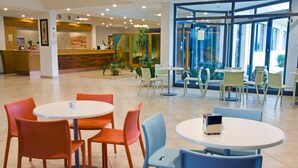 Un vestíbulo de hotel con mostrador de recepción, varias zonas de estar con mesas y sillas, y un amplio escaparate de vidrio que da al exterior.