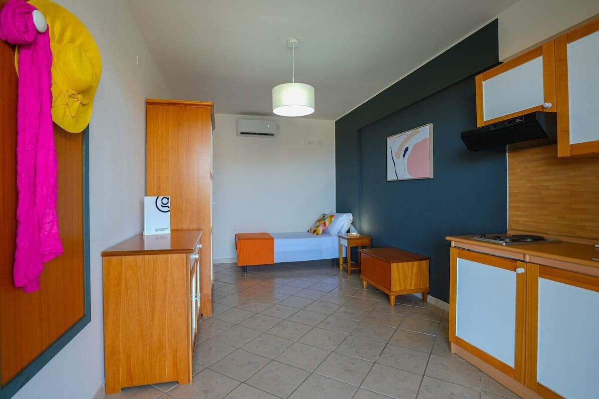 A compact room with a bed, a desk, a small table, a kitchenette, and a wall-mounted air conditioner.