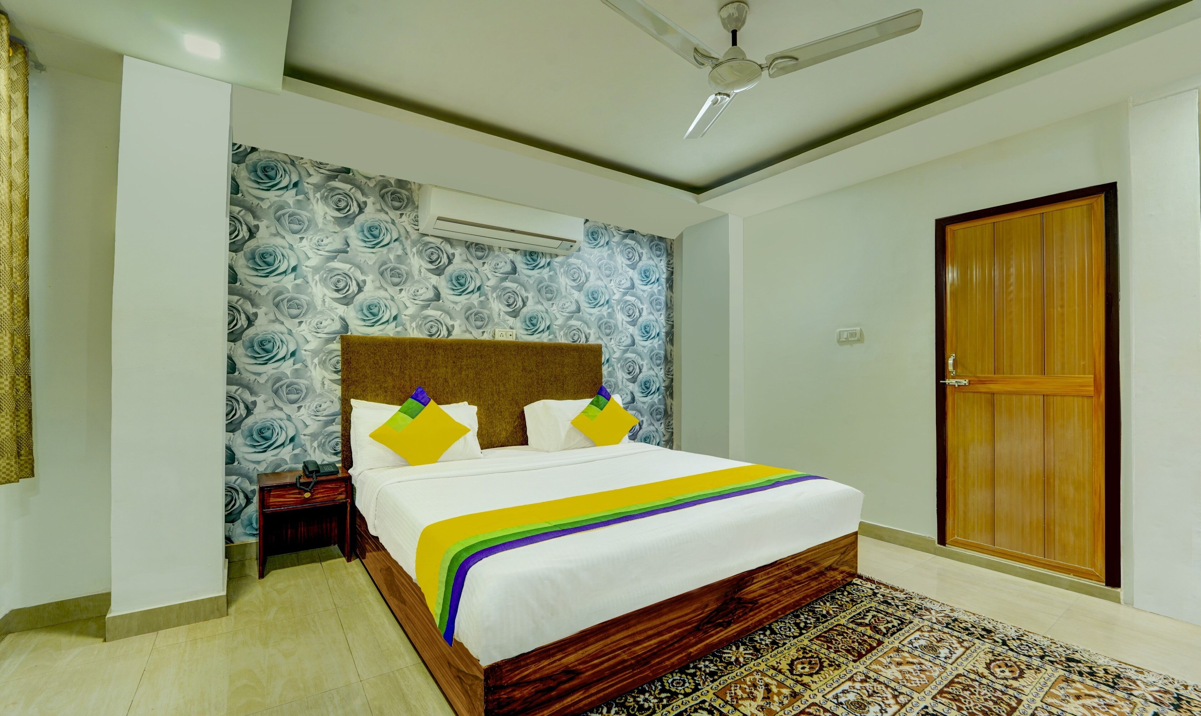 A bedroom with a bed, a ceiling fan, a patterned wall, a wooden door, and a patterned rug.