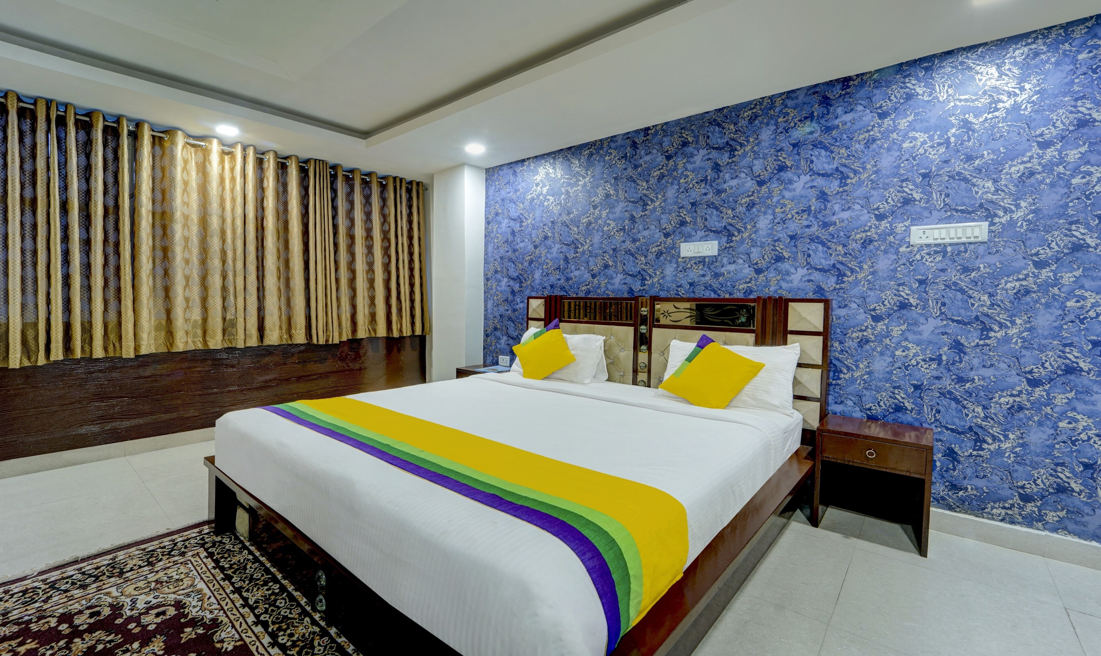Een hotelkamer met een groot bed, een nachtkastje, een muur met blauw behang met een patroon en een raam met gordijnen.