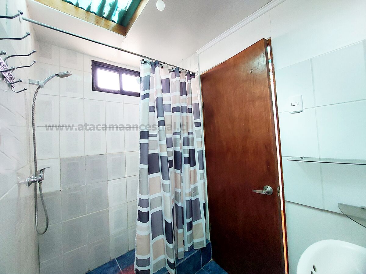 A bathroom with white tiled walls, a shower with a handheld showerhead, a wooden door, and a small window.