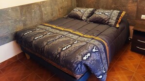 Una cama con ropa de cama y almohadas estampadas, una mesita de noche de madera con cajones, y un piso de baldosas.