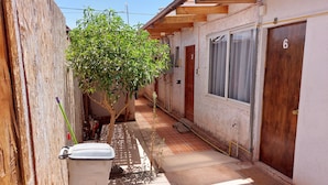 Un callejón estrecho con una puerta de madera a la izquierda, un bote de basura blanco y un árbol en el centro.