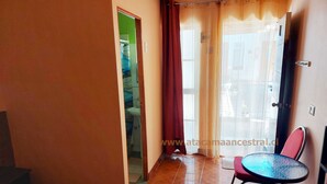 Una habitación con suelo de baldosas, una pequeña mesa redonda con sillas, una puerta que da al baño y una ventana con cortinas.