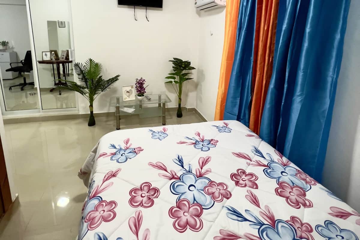 A hotel room with a bed, a TV mounted on the wall, a desk with a chair, and a window with curtains.