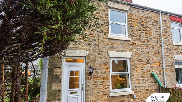 A stone house with a white door, two windows, and a small garden area with a tree. - Luke Stays -Good StationCottages (Durham)