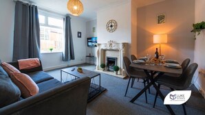 A modern living room with a sofa, a dining table set for four, a fireplace, and a television. - Luke Stays - Rokeby Street (Newcastle Upon Tyne)