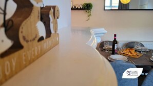 A dining area with a wooden sign reading "JOY EVERY MOMENT," a mounted TV, and a table set with wine glasses, plates, and a bottle. - Luke Stays - Rokeby Street (Newcastle Upon Tyne)