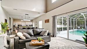 A spacious living room with a leather sofa, a dining area, and a kitchen in the background. There is a large sliding glass door leading to a pool area. - Family friendly activities for all ages! Pool, Golf, Corn-Hole, Fishing & more! (Bradenton)