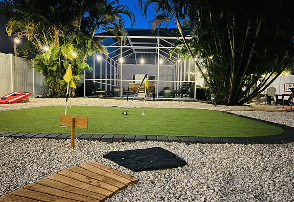 Un jardin bien éclairé avec un petit terrain d’entraînement au golf, un banc en bois et un kiosque.