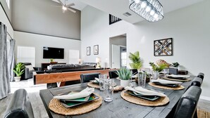 A modern dining area with a long wooden table set for a meal, a television in the background, and a ceiling fan. - Family friendly activities for all ages! Pool, Golf, Corn-Hole, Fishing & more! (Bradenton)