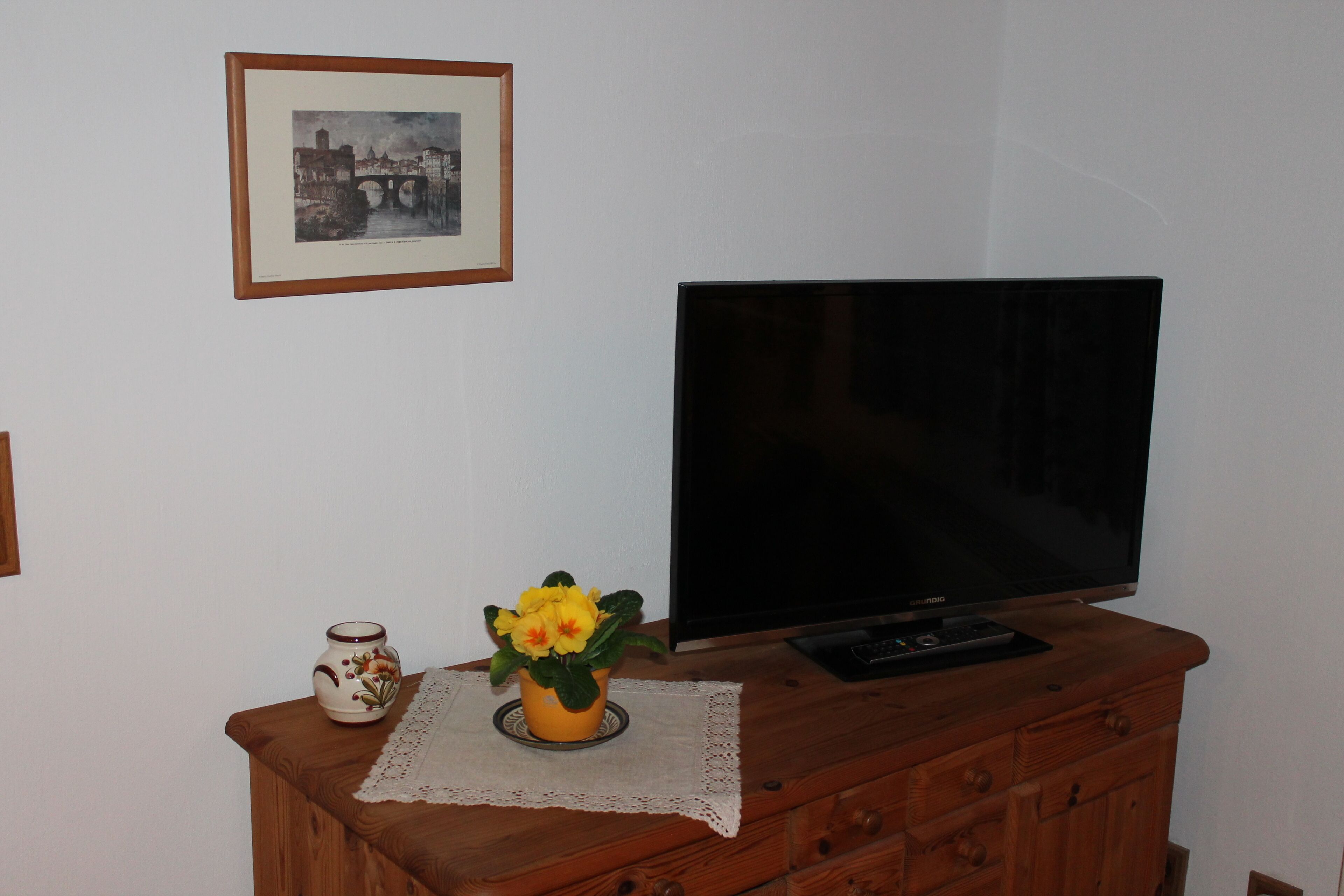 A television on a wooden dresser, a framed picture on the wall, and a vase with yellow flowers.