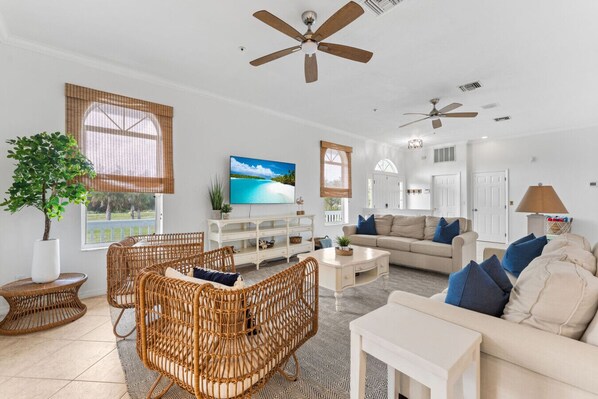 A spacious living room with wicker furniture, a flat-screen TV, and a ceiling fan.
