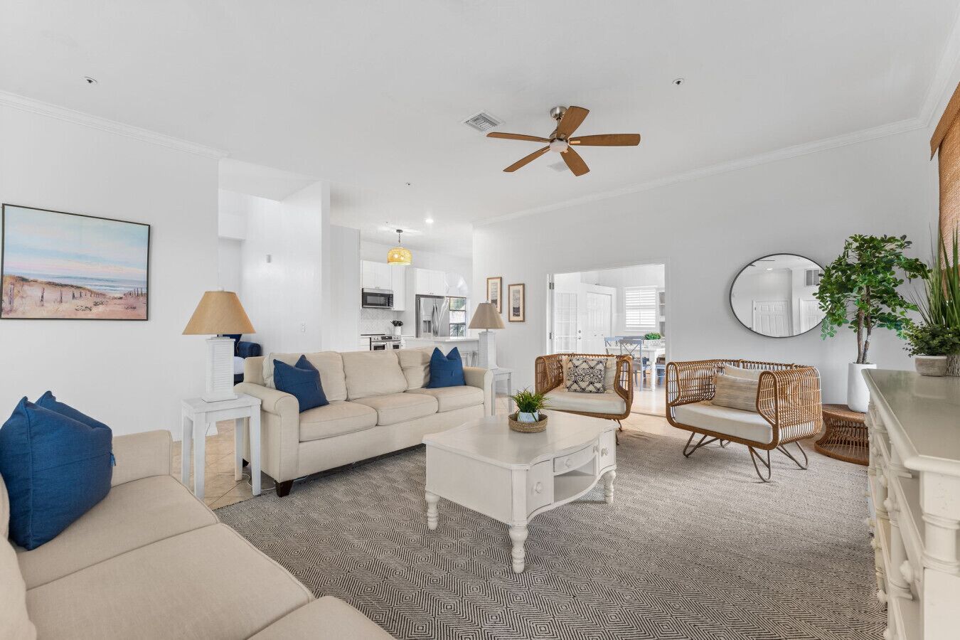 A spacious living room with a beige sofa, a white coffee table, and wicker chairs. A ceiling fan and a kitchen are visible in the background.
