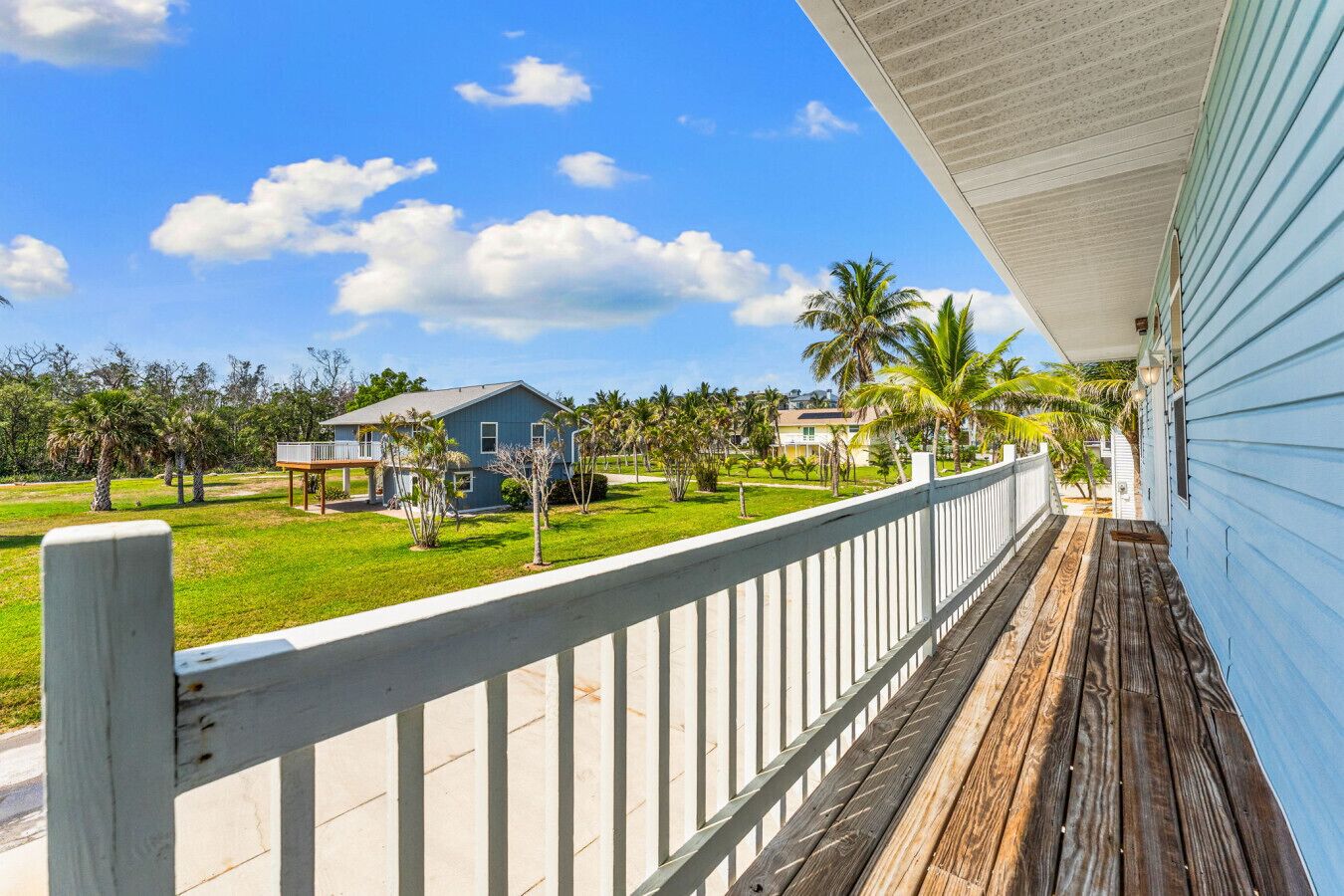 Une terrasse en bois avec une balustrade blanche, donnant sur une zone verdoyante parsemée de palmiers et sur une maison bleue en arrière-plan.