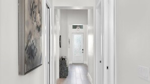 A narrow hallway with a white door at the end, a light fixture above it, and a framed abstract painting on the left wall. - Brand New Luxury Pool & Spa Home Near Beach! (Naples)