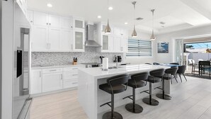A modern kitchen with white cabinets, a marble countertop, and a central island with bar stools. - Brand New Luxury Pool & Spa Home Near Beach! (Naples)