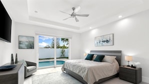 A modern bedroom with a large bed, a ceiling fan, a nightstand with a lamp, and a view of a swimming pool through glass doors. - Brand New Luxury Pool & Spa Home Near Beach! (Naples)