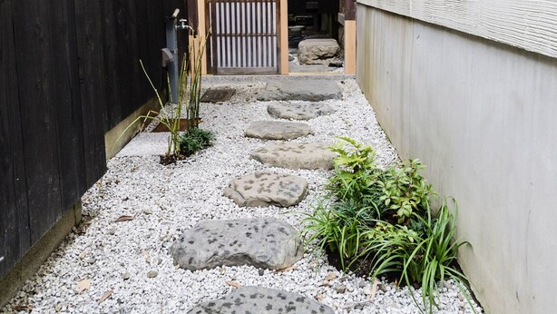 Un étroit sentier mène à une entrée de style japonais traditionnel, dotée d’une porte coulissante.
