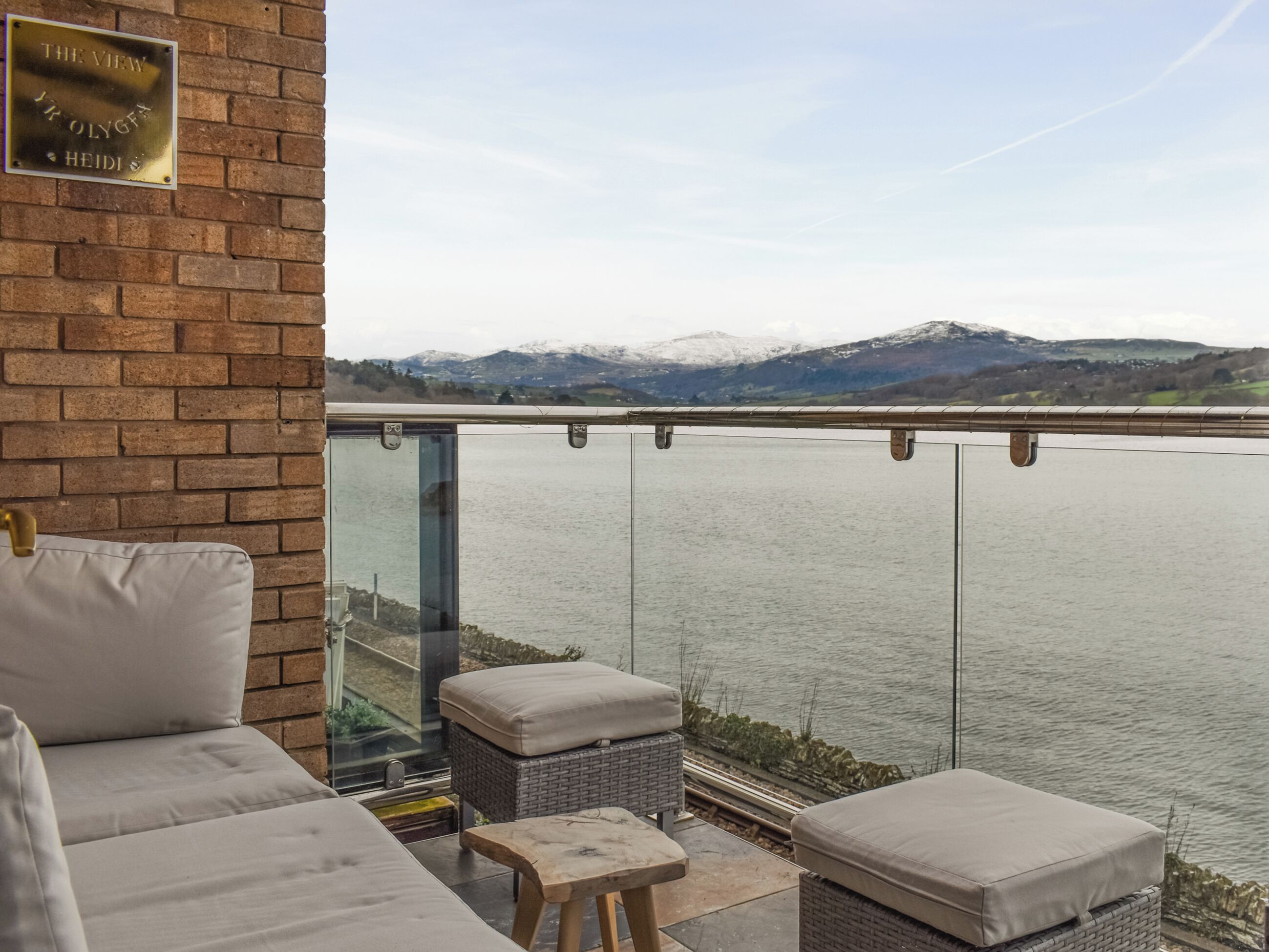 A balcony with a view of a river and mountains, furnished with a sofa and ottoman, and a plaque on the wall.