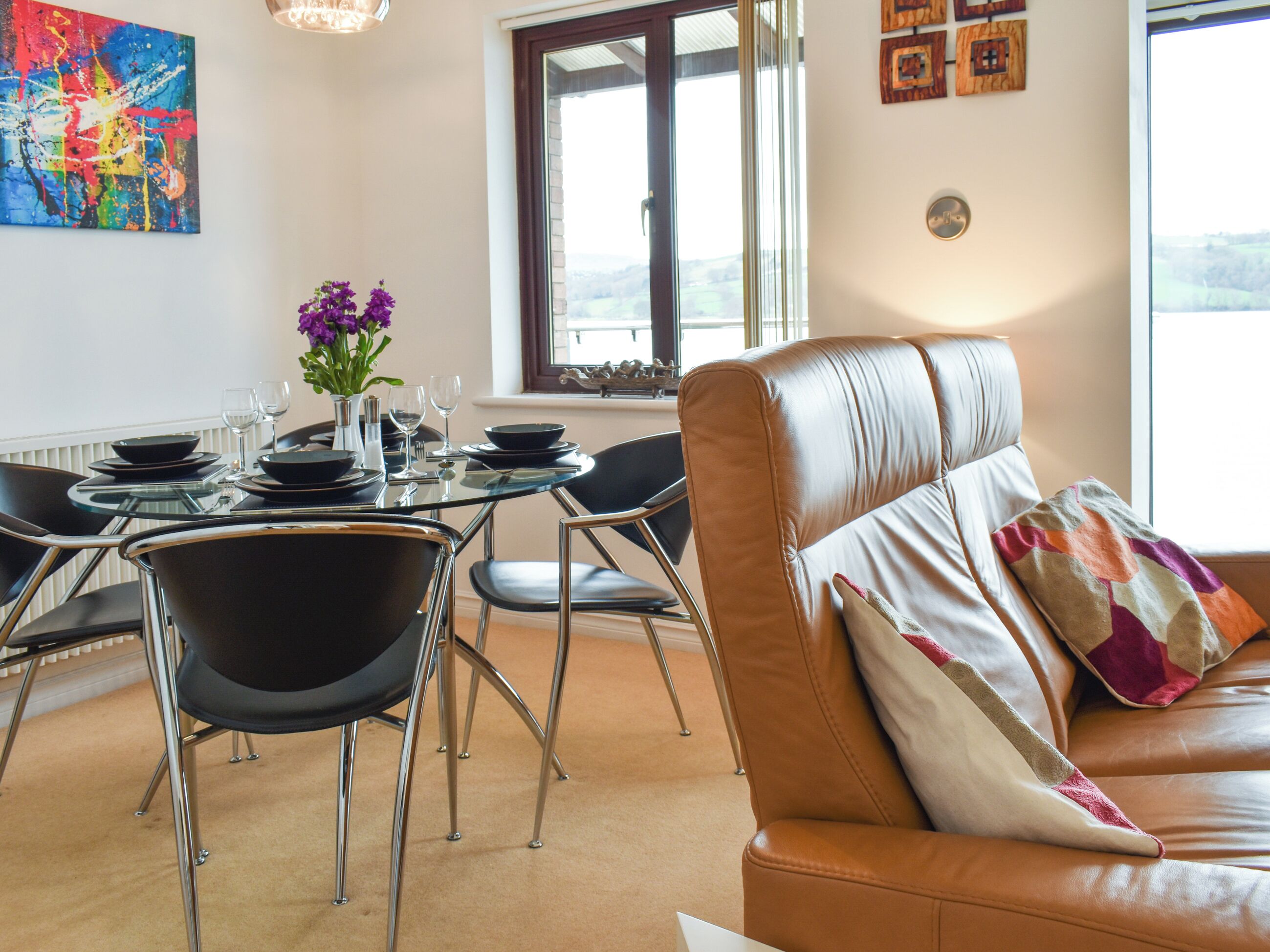 A dining area with a glass table, black chairs, and a leather couch with a colorful pillow.