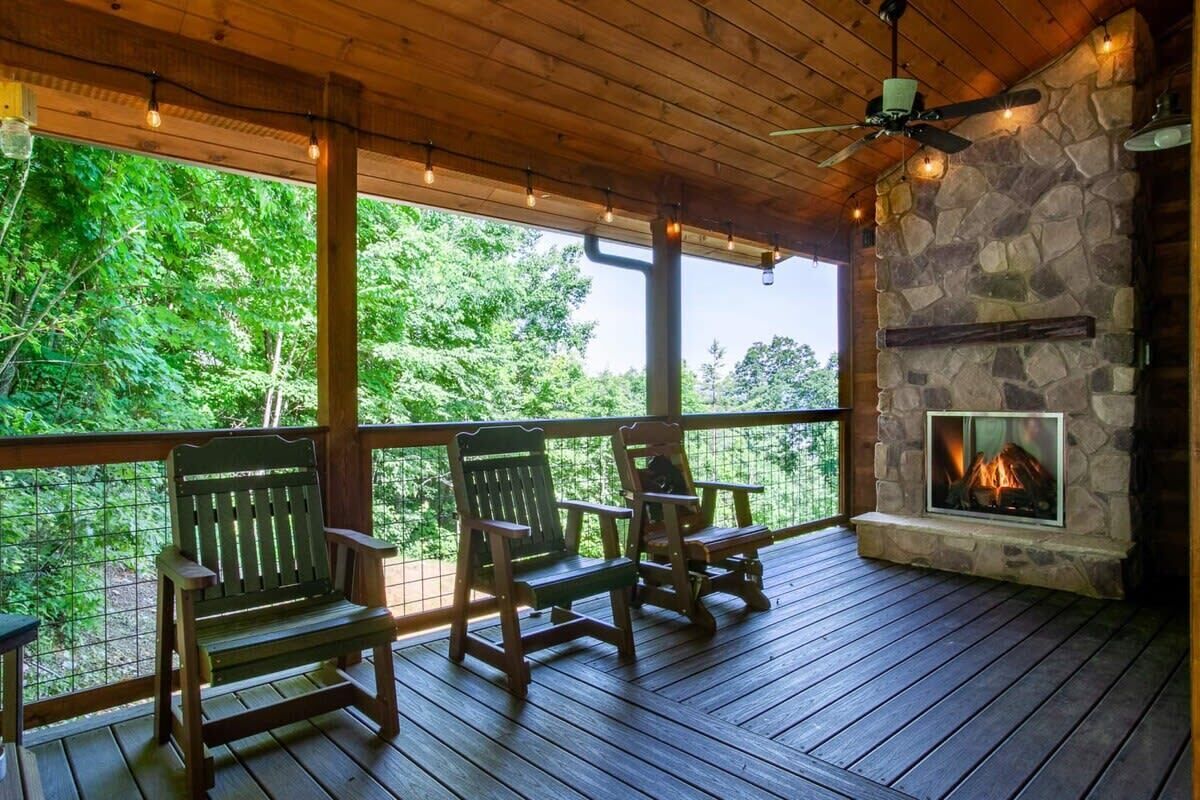 Eine Holzterrasse mit zwei Adirondack-Stühlen, einem Kamin aus Stein und Blick ins Waldgebiet.