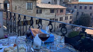 A table with a colorful tablecloth, a glass of red wine, a glass of water, and a plate of food on a balcony overlooking historic buildings and a tower. - The Penthouse of Dreams (san gimignano)