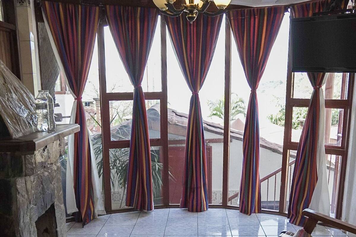 Ein Hotelzimmer mit einem Bett, einem grünen Handtuch, einem hölzernen Schaukelstuhl, einem Kamin aus Stein und großen Fenstern mit gestreiften Vorhängen.