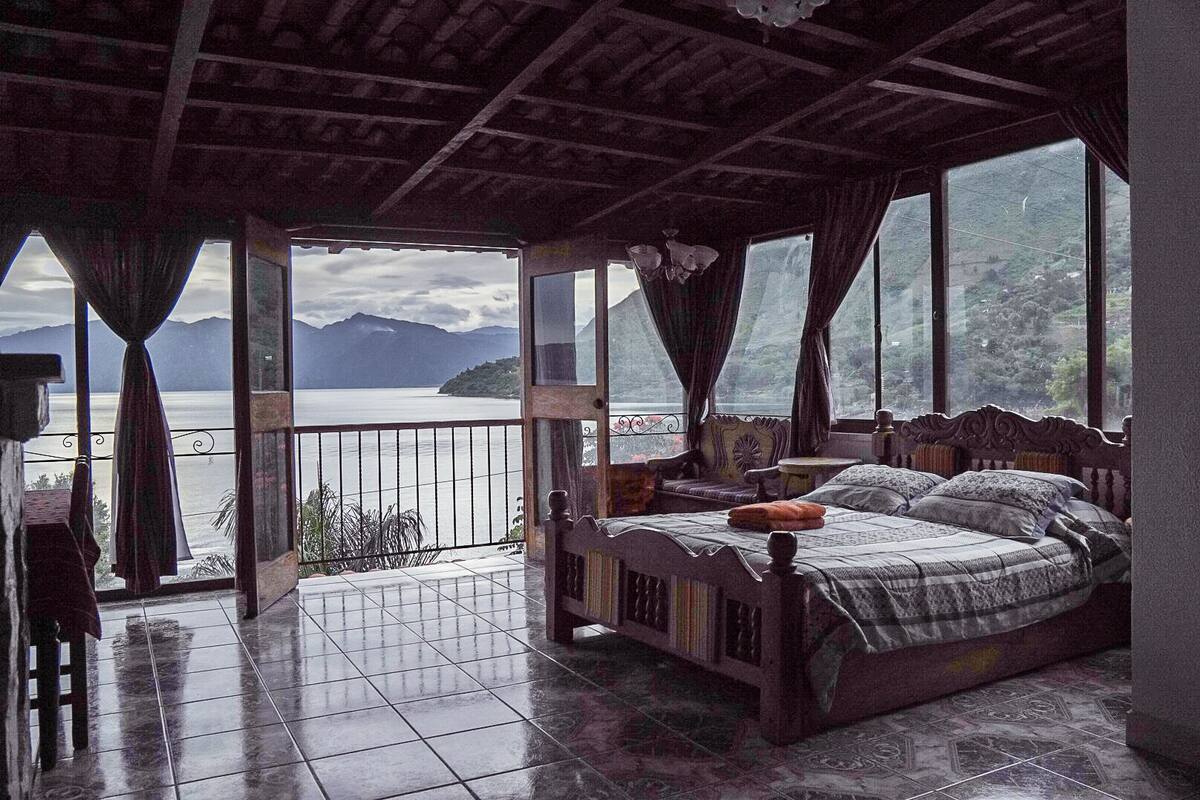 Ein Schlafzimmer mit Bett, kleinem Tisch und Stuhl. Es besitzt einen Balkon mit Blick auf einen See und Berge.