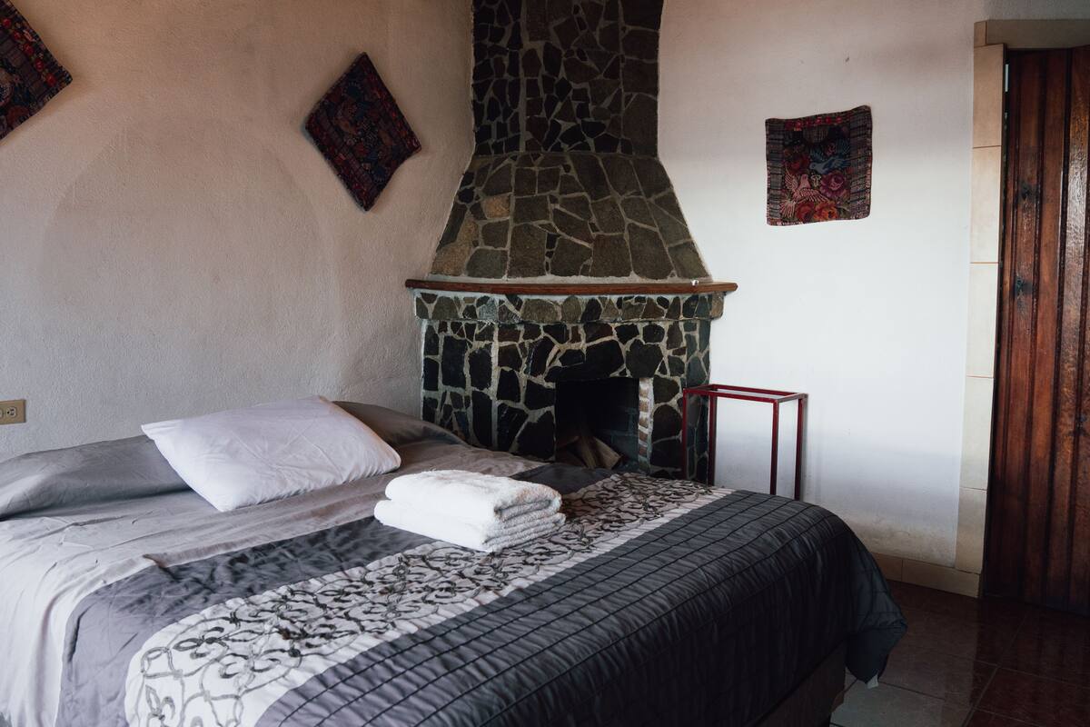 Ein Schlafzimmer mit Kamin aus Stein, einem ordentlich bezogenen Bett und einem kleinen Tisch neben dem Bett.