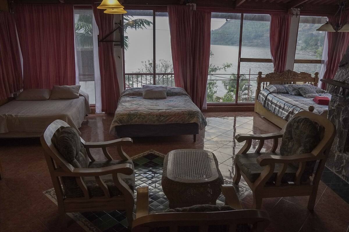 Ein Zimmer mit zwei Betten, einem Schlafsofa und einem Esstisch. Große Fenster bieten Blick auf ein Gewässer und Hügel.