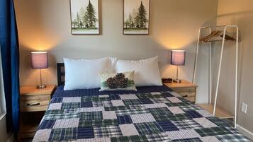 A neatly made bed with a plaid quilt, two white pillows, and a pinecone decoration. There are two framed pictures on the wall, a bedside lamp, and a coat rack.