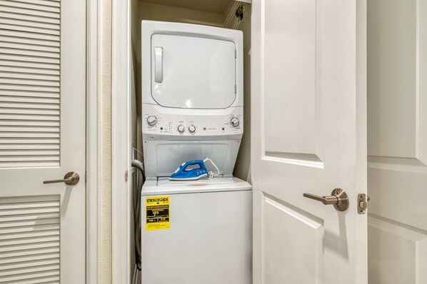 Une buanderie équipée d’un lave-linge et d’un sèche-linge à chargement frontal, d’un fer à repasser bleu et d’un autocollant jaune indiquant le niveau de performance énergétique.