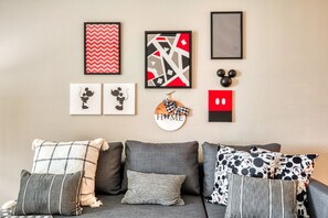 Sala de estar com sofá cinzento, vários almofadas e obras de arte nas paredes, incluindo silhuetas do Mickey Mouse e designs abstratos.