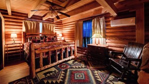 Une chambre dans un chalet en rondins, avec un lit en bois, un fauteuil à bascule, un tapis à motifs et un ventilateur de plafond.