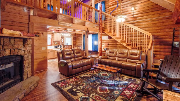 L’intérieur d’une cabane en rondins, avec une cheminée en pierre, des canapés en cuir, un escalier en bois et une vue sur la cuisine.